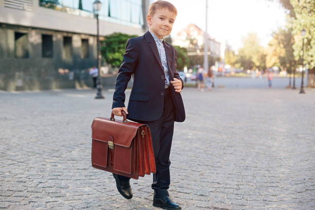 Concept future businessman. Business meeting. Low angle of future businessman in formal wear walking around the street carrying a braun briefcase and holding jacket with arm, urban background.